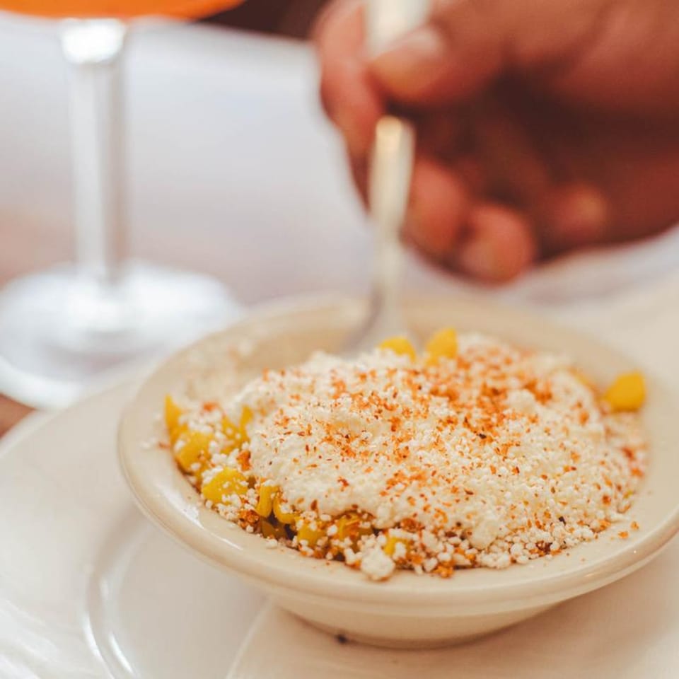 A bowl of Mexican street corn (Esquites) with cheese and chili powder, being eaten with a spoon.