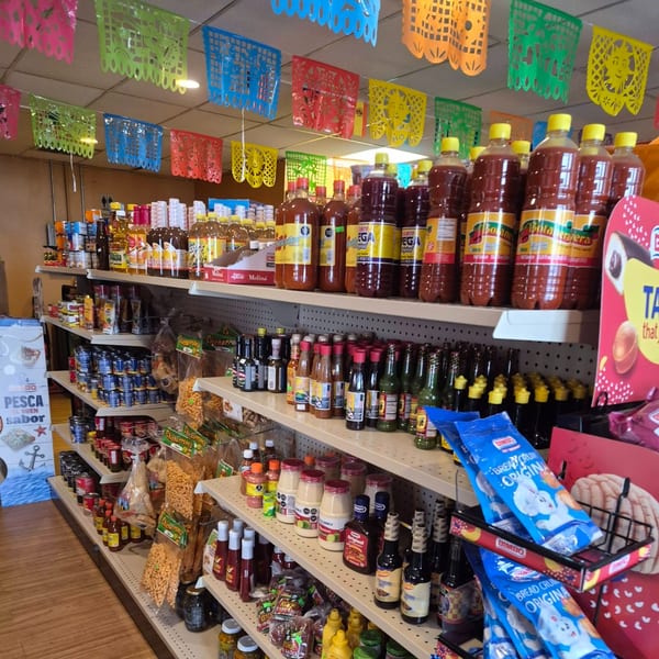 Shelves filled with various Mexican groceries, sauces, and snacks under colorful papel picado decorations.