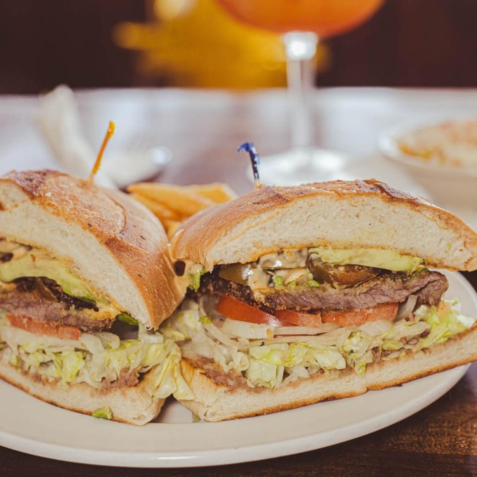 A large Mexican torta sandwich, cut in half, filled with meat, lettuce, tomato, and avocado.