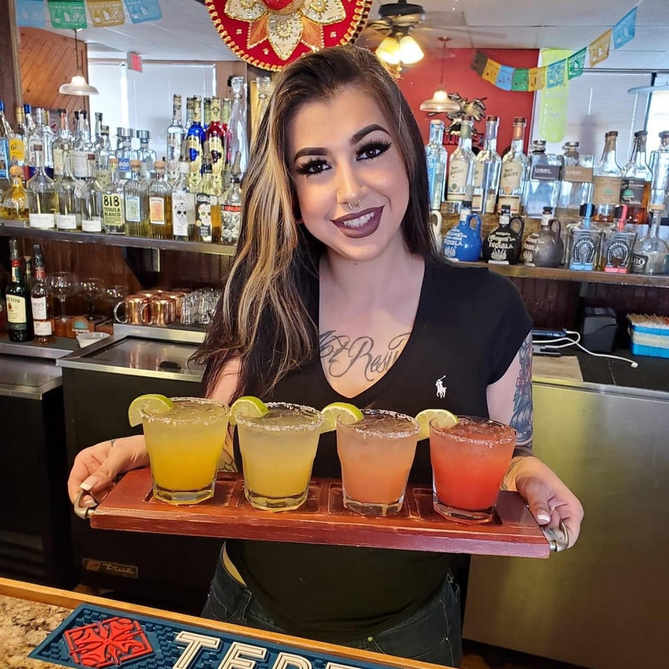 Bartender at LA Taco Cantina holding a tray with four colorful margaritas, with a tequila bar in the background.