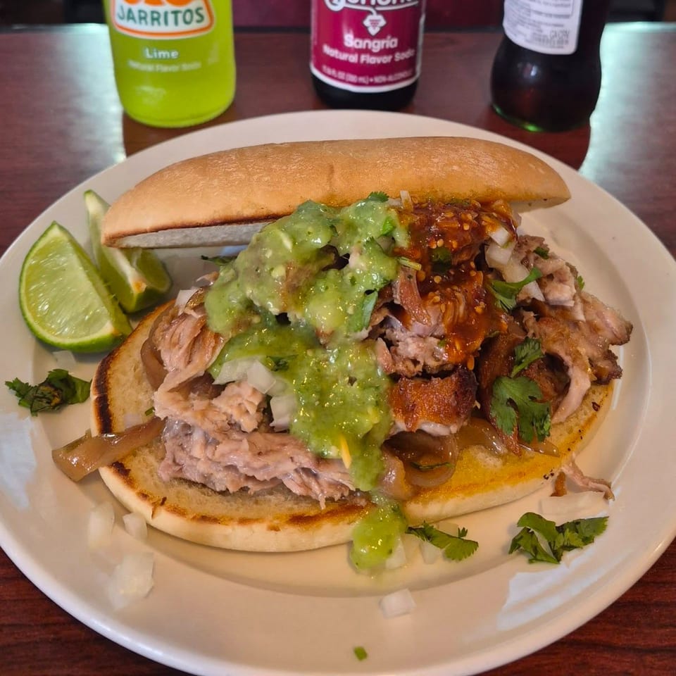 A Mexican torta with meat, guacamole, and salsa, served with lime wedges and Jarritos sodas.