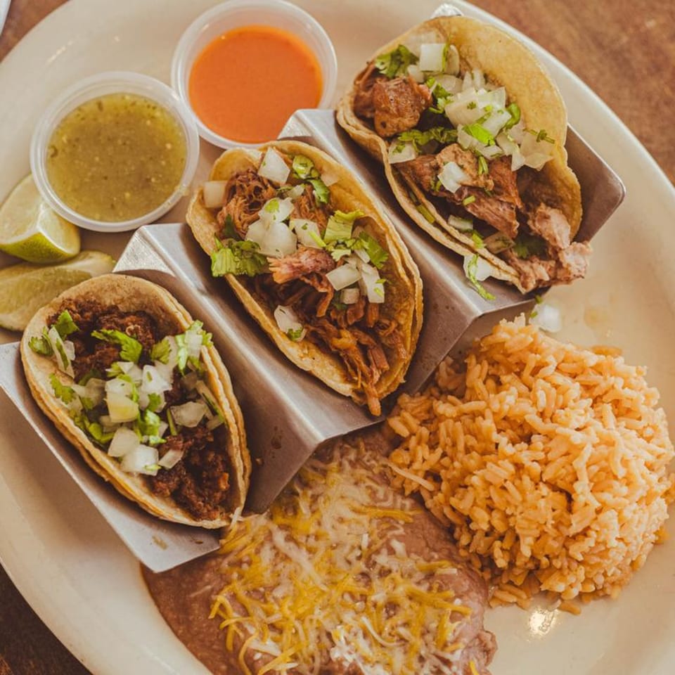 Three LA Taco Cantina tacos with rice, beans, lime, and two sauces on a white plate.