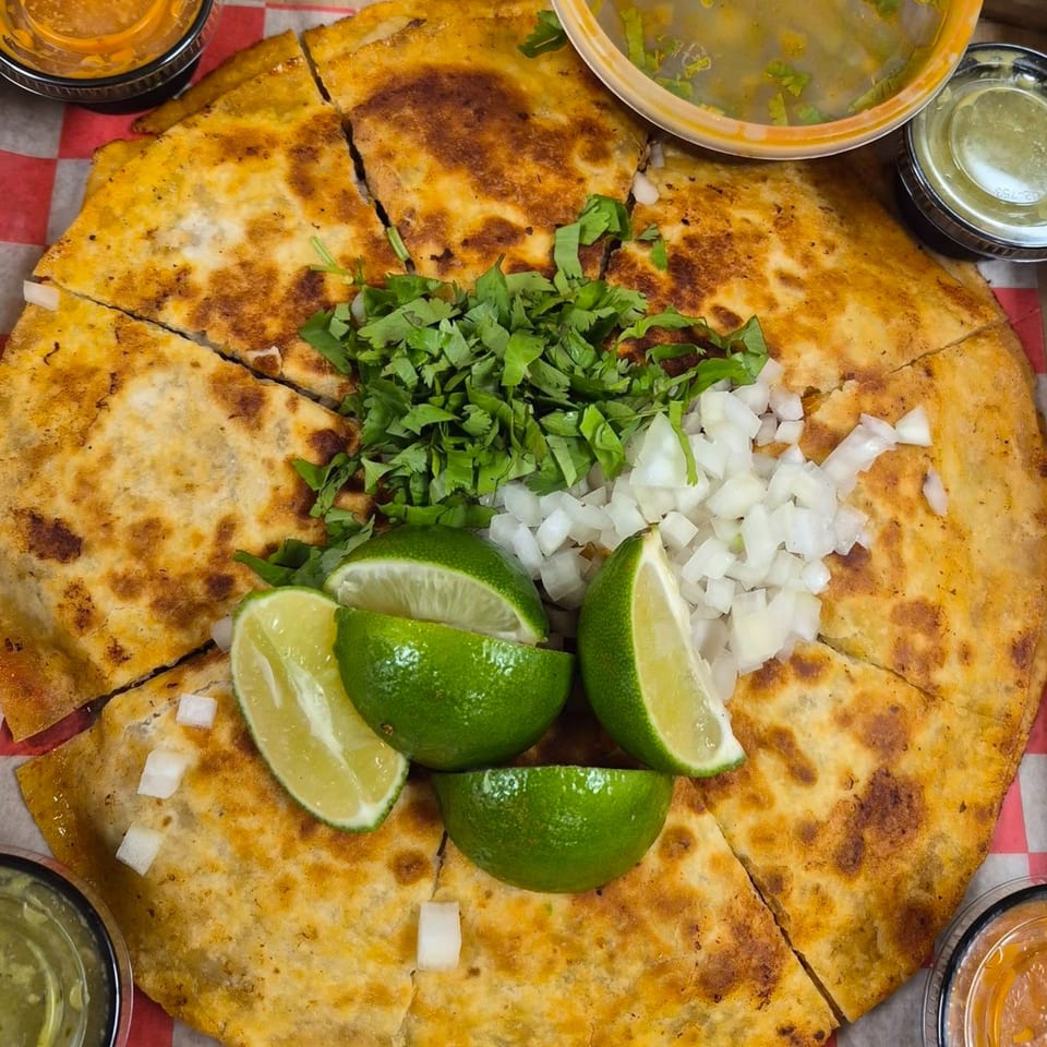 A large, sliced quesadilla topped with cilantro, diced onions, and lime wedges, with dipping sauces.
