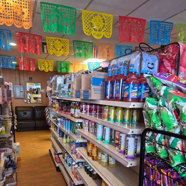 Colorful papel picado banners hang above shelves stocked with Mexican groceries and beverages in a store.