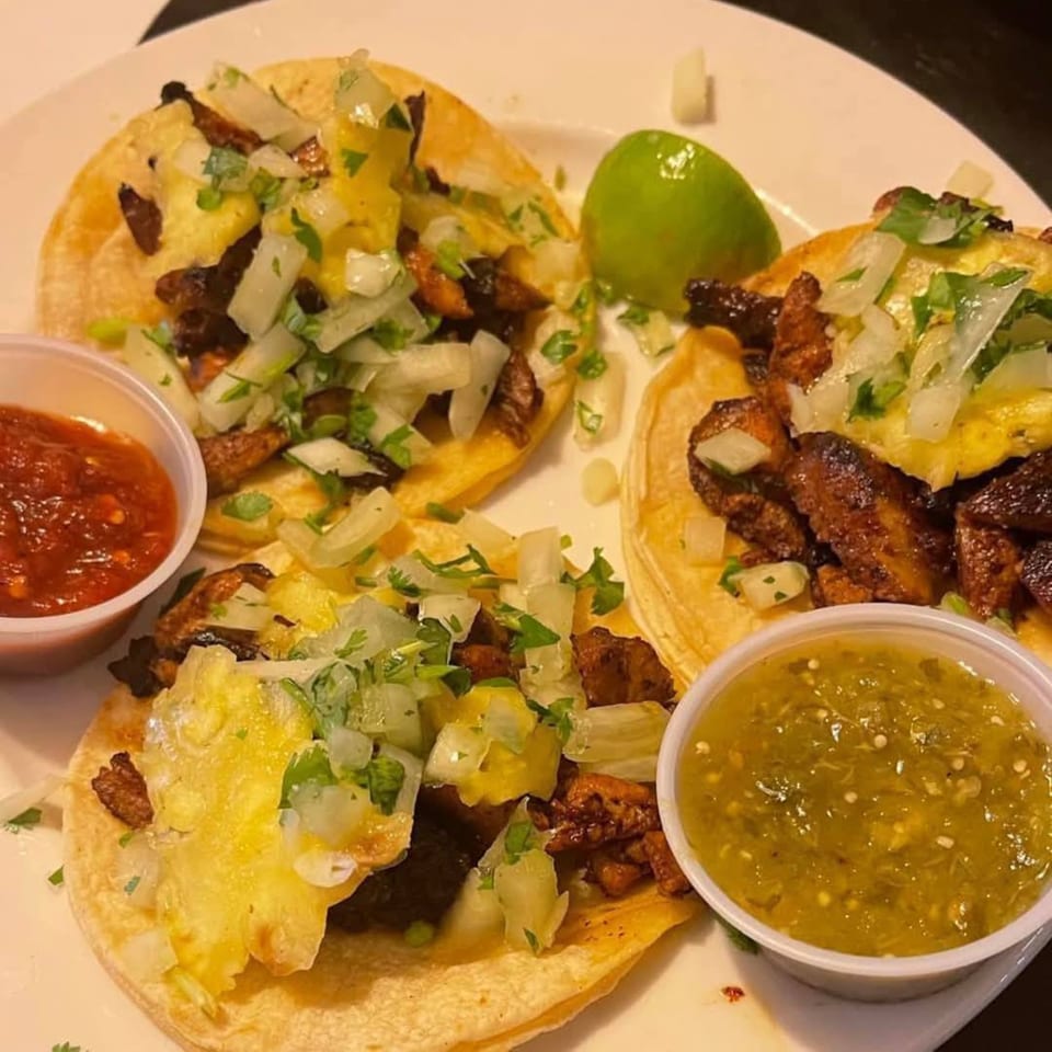 Three al pastor tacos with pineapple, onions, cilantro, and two salsas on a white plate.